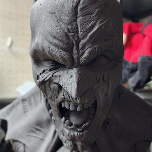 May include: A dark gray, unpainted resin bust of a character wearing a cowl with pointed ears. The face features a weathered, textured surface with a snarling expression, exposed teeth, and deep-set eyes. The base includes a bat-shaped emblem.