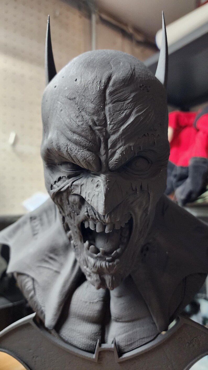 May include: A dark gray, unpainted resin bust of a character wearing a cowl with pointed ears. The face features a weathered, textured surface with a snarling expression, exposed teeth, and deep-set eyes. The base includes a bat-shaped emblem.