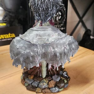 Op de afbeelding: Een 3D-geprint borstbeeld van een figuur met donker, gestructureerd haar, gekleed in een gehavende, verweerde grijze mantel over een donkerbruin kledingstuk. Het figuur staat op een voetstuk van onregelmatige, bruin- en grijskleurige stenen.