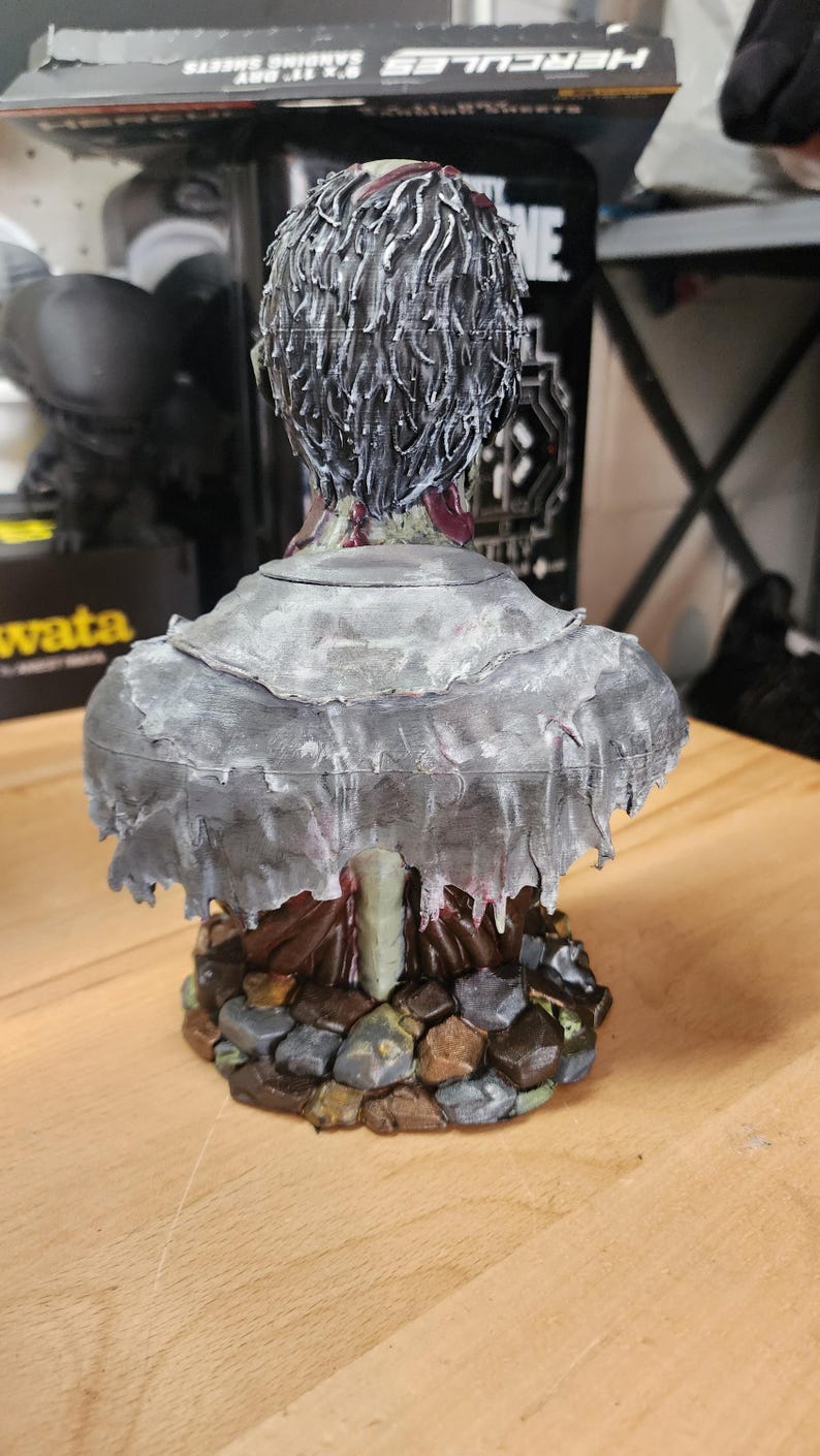 Op de afbeelding: Een 3D-geprint borstbeeld van een figuur met donker, gestructureerd haar, gekleed in een gehavende, verweerde grijze mantel over een donkerbruin kledingstuk. Het figuur staat op een voetstuk van onregelmatige, bruin- en grijskleurige stenen.