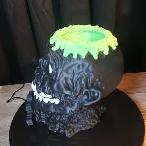 May include: A black skull-shaped container with a textured surface, featuring a bright green drip-like rim at the top. The skull has white painted teeth and eye details, with a hollow interior, presented as a decorative object for storage.
