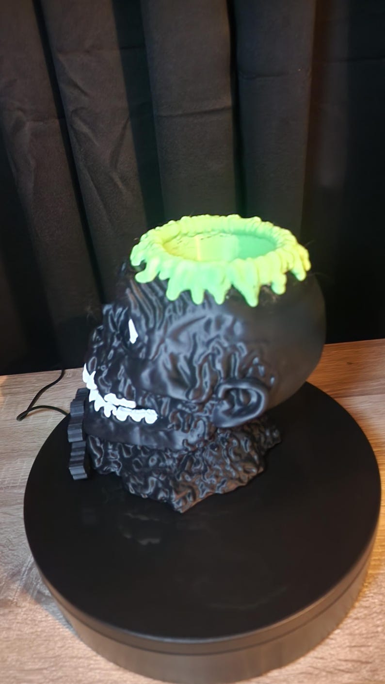 May include: A black skull-shaped container with a textured surface, featuring a bright green drip-like rim at the top. The skull has white painted teeth and eye details, with a hollow interior, presented as a decorative object for storage.