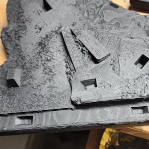 May include: A black 3D printed terrain tile featuring a textured rocky surface with recessed square openings and raised geometric structures. The piece displays visible layer lines from the printing process and includes a separate black component.