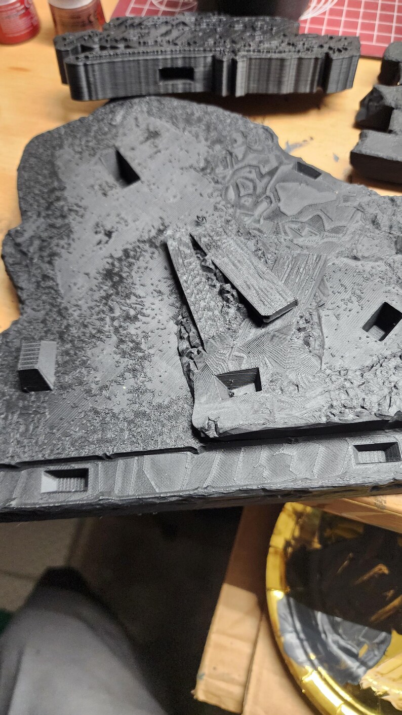 May include: A black 3D printed terrain tile featuring a textured rocky surface with recessed square openings and raised geometric structures. The piece displays visible layer lines from the printing process and includes a separate black component.