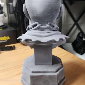 May include: A gray 3D printed bust of a bald figure viewed from the back. The figure wears a collar with a chain link detail around the neck, resting on a pedestal base. The surface has a matte finish with visible layer lines from the printing process.