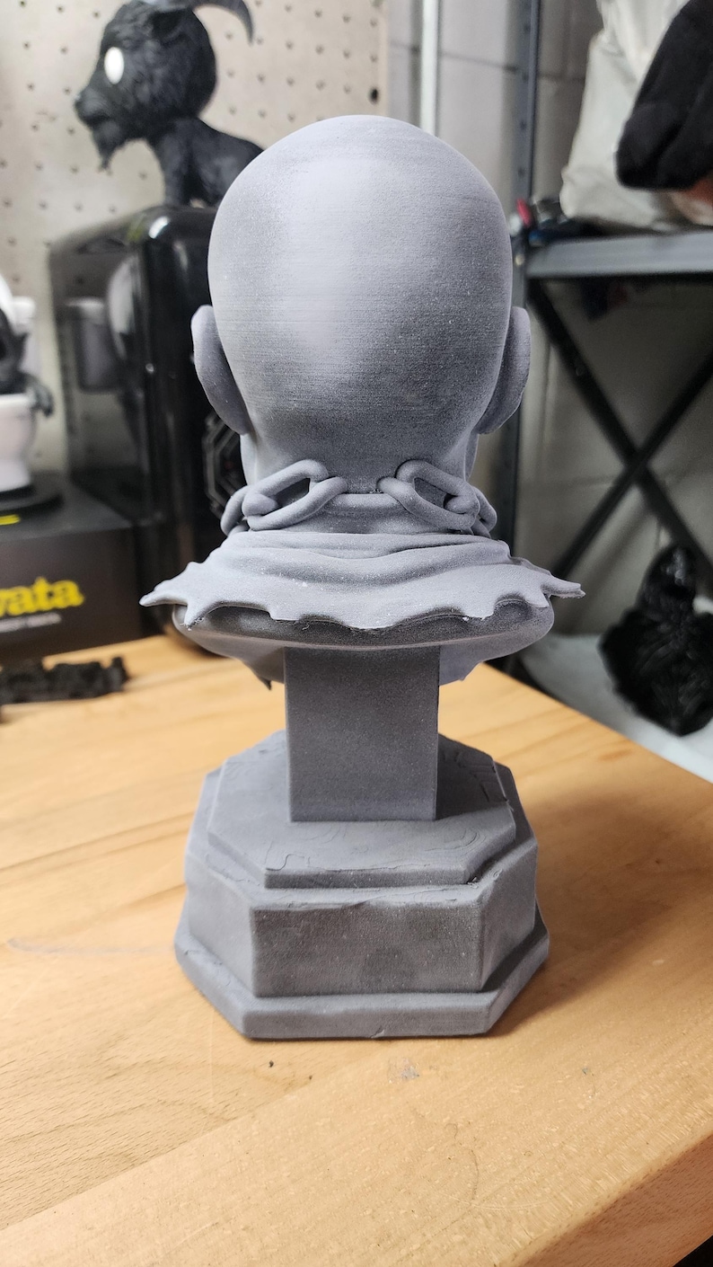 May include: A gray 3D printed bust of a bald figure viewed from the back. The figure wears a collar with a chain link detail around the neck, resting on a pedestal base. The surface has a matte finish with visible layer lines from the printing process.