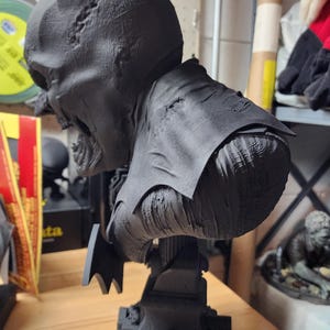 May include: A matte black 3D printed bust of a character with pointed ears and a tattered cape, featuring a textured, weathered surface. The figure is mounted on a square base designed to resemble a tombstone with a small arched door detail.