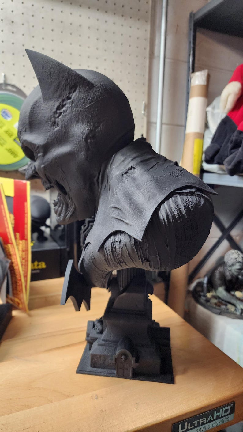 May include: A matte black 3D printed bust of a character with pointed ears and a tattered cape, featuring a textured, weathered surface. The figure is mounted on a square base designed to resemble a tombstone with a small arched door detail.