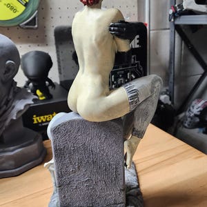 Puede incluir: Una figura con cabello corto de color marr&oacute;n rojizo, sentada sobre una l&aacute;pida gris texturizada. La figura tiene piel p&aacute;lida, usa guantes negros y tiene mallas grises estampadas en la parte inferior de las piernas. La base es de piedra gris rugosa.
