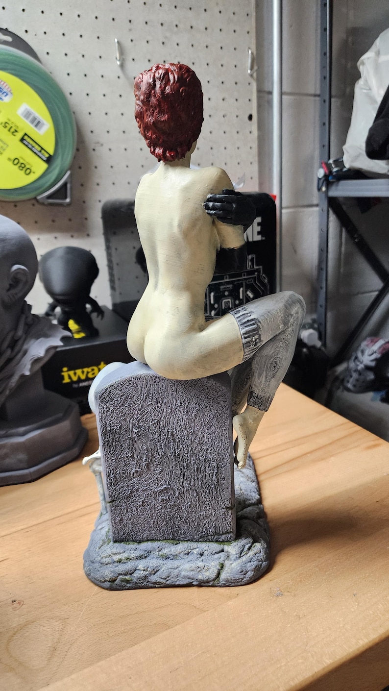 Puede incluir: Una figura con cabello corto de color marr&oacute;n rojizo, sentada sobre una l&aacute;pida gris texturizada. La figura tiene piel p&aacute;lida, usa guantes negros y tiene mallas grises estampadas en la parte inferior de las piernas. La base es de piedra gris rugosa.