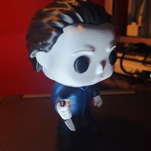 May include: A stylized vinyl figure of a character with a white mask, black hair, and solid black eyes. The figure wears a dark blue jumpsuit and holds a small silver-colored knife in its right hand. The design features a large head and small body.