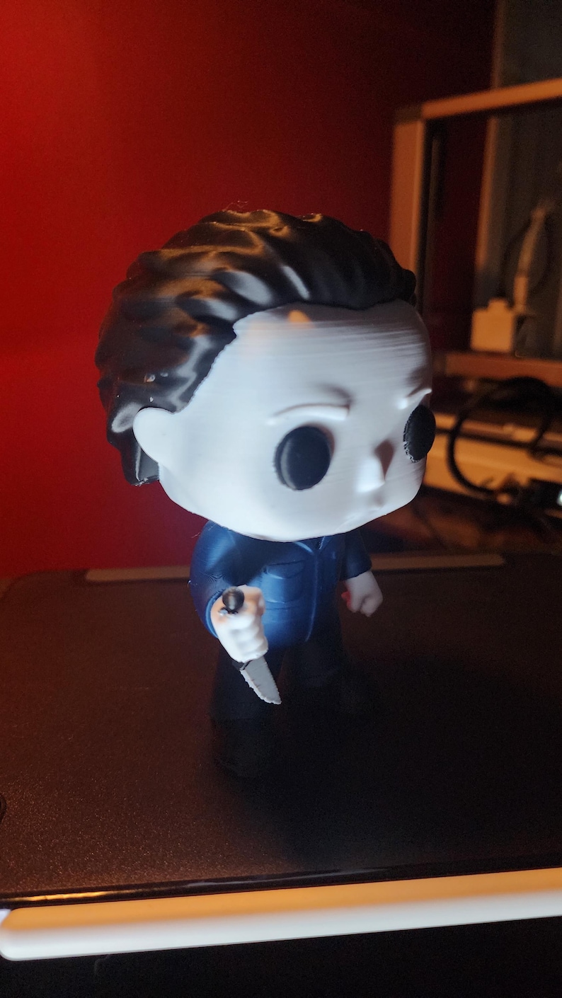 May include: A stylized vinyl figure of a character with a white mask, black hair, and solid black eyes. The figure wears a dark blue jumpsuit and holds a small silver-colored knife in its right hand. The design features a large head and small body.