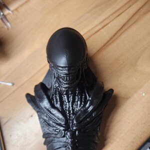 May include: A matte black bust of a creature with an elongated, smooth cranium, exposed rib-like structures on the chest, and a detailed jawline featuring visible teeth. The sculpture displays intricate, organic textures across the neck and torso.
