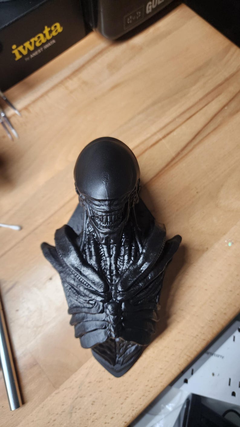 May include: A matte black bust of a creature with an elongated, smooth cranium, exposed rib-like structures on the chest, and a detailed jawline featuring visible teeth. The sculpture displays intricate, organic textures across the neck and torso.