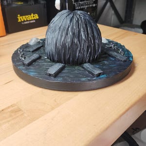 May include: A circular display base featuring a dark, textured water surface with protruding wooden planks and a central, rounded, dark gray mass with vertical hair-like detailing. The base has a painted finish with blue and black highlights.