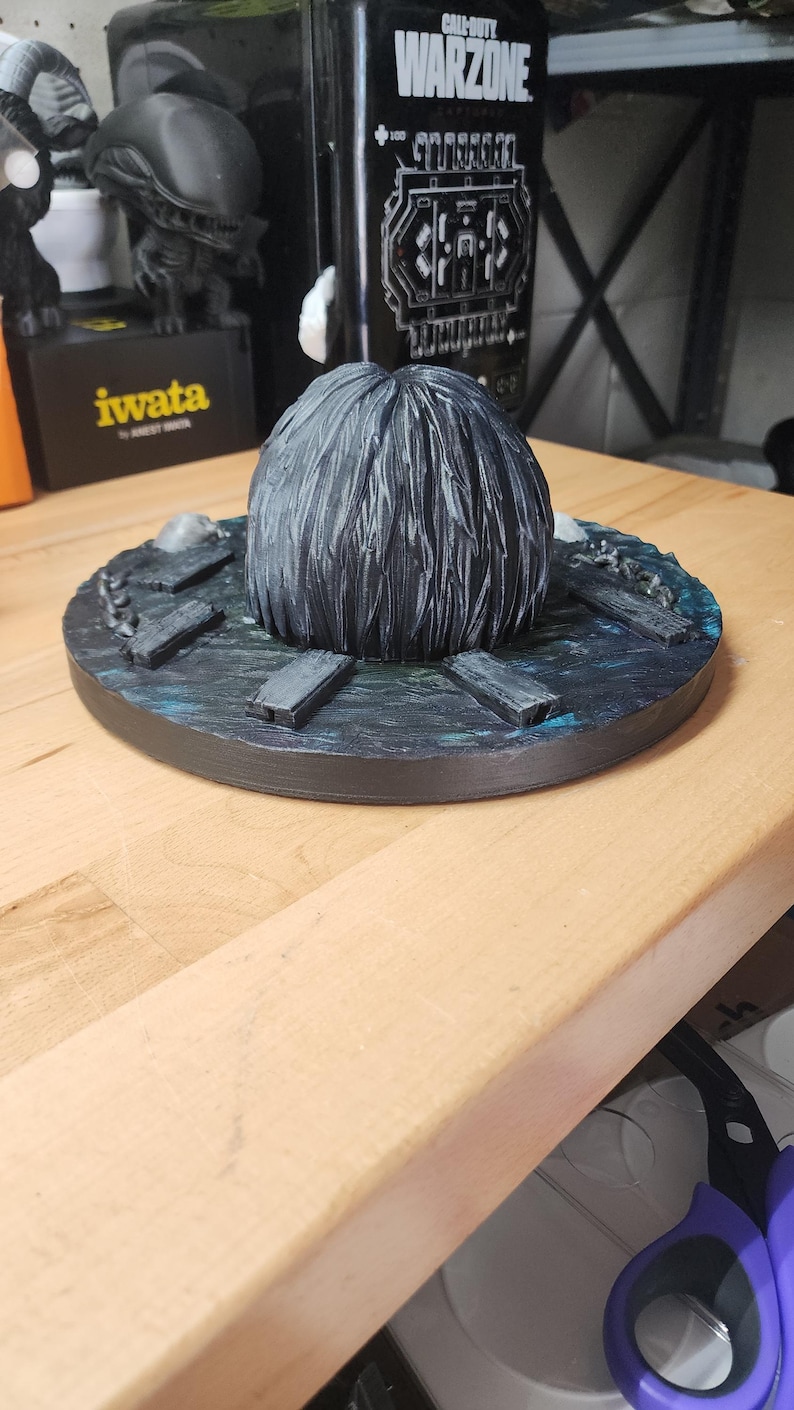 May include: A circular display base featuring a dark, textured water surface with protruding wooden planks and a central, rounded, dark gray mass with vertical hair-like detailing. The base has a painted finish with blue and black highlights.