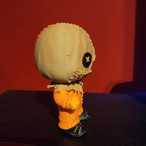 May include: A small collectible figurine featuring a round, beige burlap-textured head with a single black button eye and stitched mouth details. The figure wears a bright orange jumpsuit with frayed collar edges and black shoes with yellow accents.