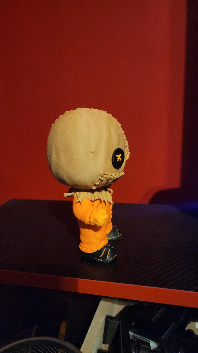 May include: A small collectible figurine featuring a round, beige burlap-textured head with a single black button eye and stitched mouth details. The figure wears a bright orange jumpsuit with frayed collar edges and black shoes with yellow accents.