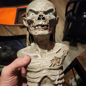 May include: A beige, skeletal bust of the Crypt Keeper character with bright blue eyes, exposed ribs, and weathered skin texture. The figure is mounted on a brown rectangular base featuring the embossed white text TALES FROM THE CRYPT.
