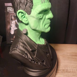 May include: A three-dimensional bust of Frankenstein's monster featuring a bright green face, dark black hair, and a black jacket. The figure has a prominent forehead, a bolt on the neck, and visible facial features. The text FRA is visible on a box.