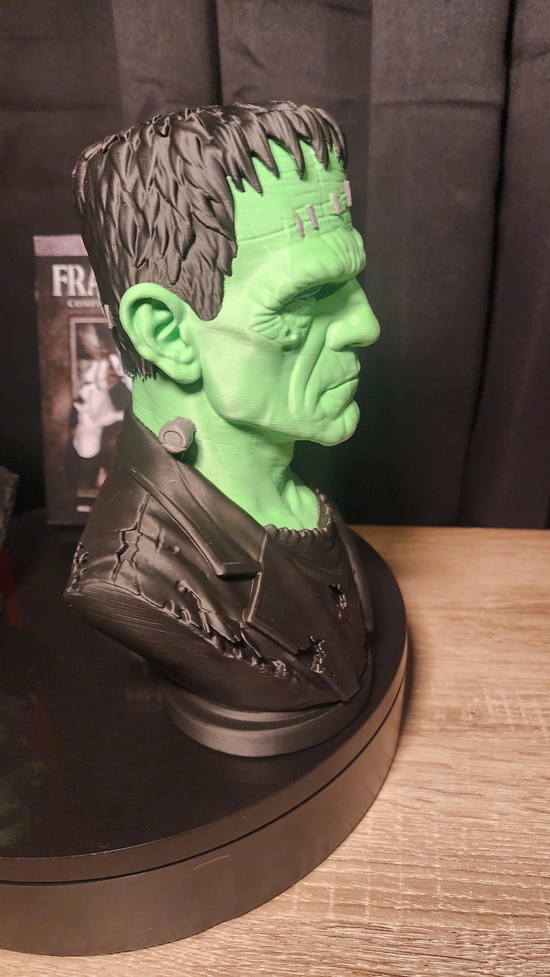 May include: A three-dimensional bust of Frankenstein's monster featuring a bright green face, dark black hair, and a black jacket. The figure has a prominent forehead, a bolt on the neck, and visible facial features. The text FRA is visible on a box.