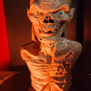 May include: A grey, skeletal bust of the Crypt Keeper character with sunken eyes, a wide grin, and textured skin. The figure is mounted on a rectangular base featuring the embossed text "TALES FROM THE CRYPT" in a metallic finish, illuminated by warm light.