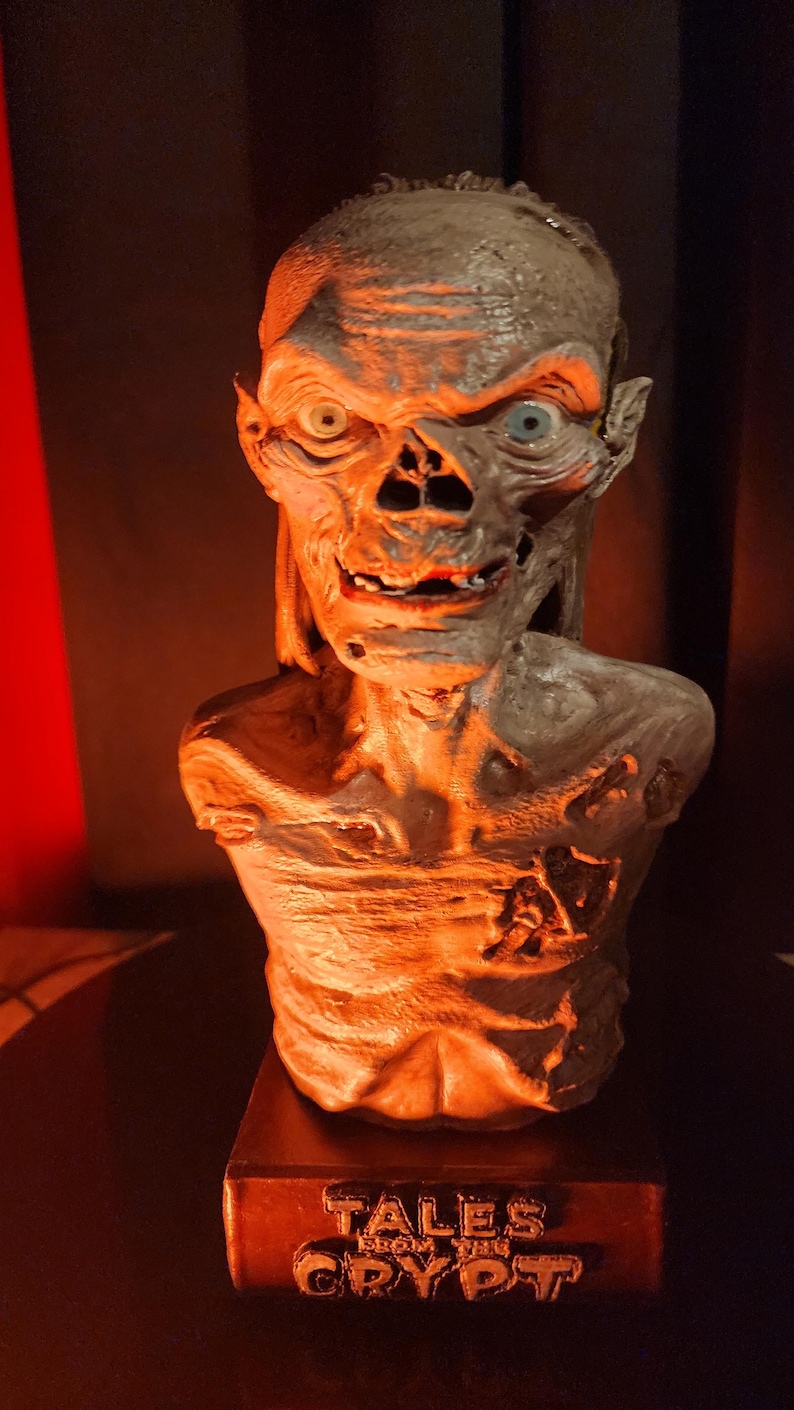 May include: A grey, skeletal bust of the Crypt Keeper character with sunken eyes, a wide grin, and textured skin. The figure is mounted on a rectangular base featuring the embossed text "TALES FROM THE CRYPT" in a metallic finish, illuminated by warm light.