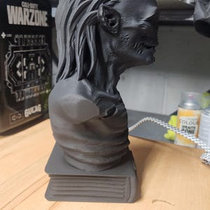 May include: A matte black 3D printed bust of a creature with long hair and textured skin, positioned on a rectangular base that resembles a closed book. The figure is shown in profile, displaying detailed facial features and a slender neck.