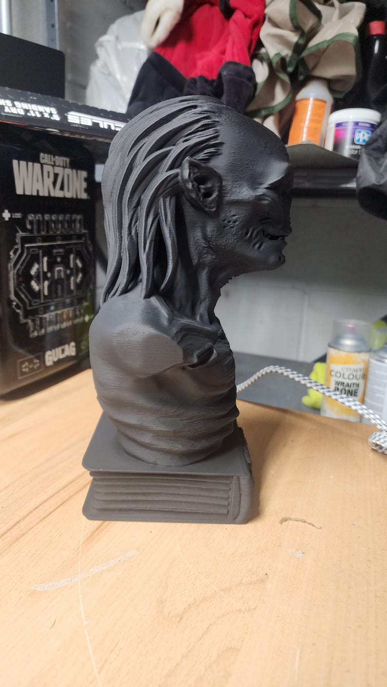 May include: A matte black 3D printed bust of a creature with long hair and textured skin, positioned on a rectangular base that resembles a closed book. The figure is shown in profile, displaying detailed facial features and a slender neck.
