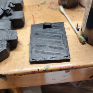 May include: A rectangular black 3D printed component featuring a series of parallel diagonal ridges across its surface and a small raised tab at the top edge. The object has a textured finish consistent with additive manufacturing processes.
