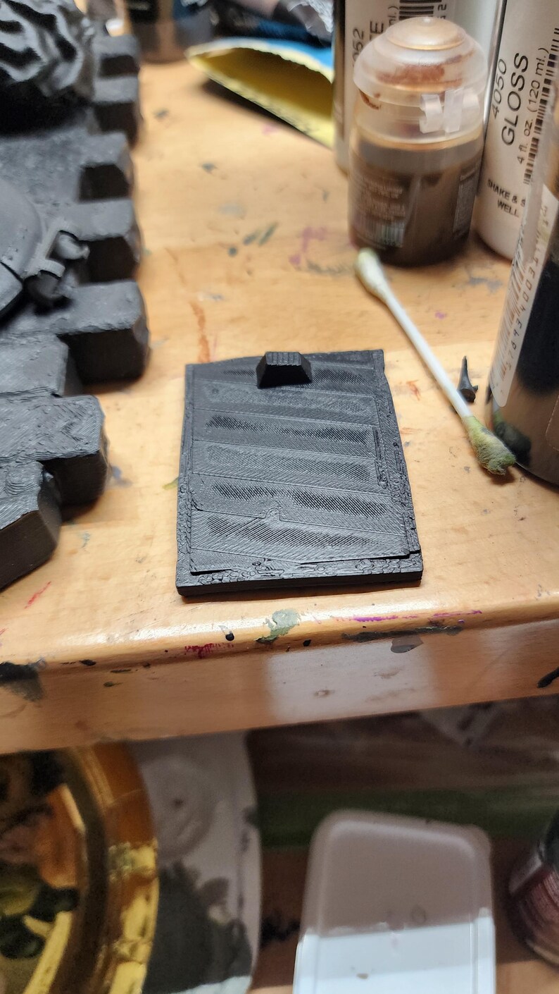 May include: A rectangular black 3D printed component featuring a series of parallel diagonal ridges across its surface and a small raised tab at the top edge. The object has a textured finish consistent with additive manufacturing processes.