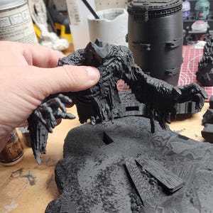 May include: A black 3D printed miniature figure of a monstrous creature with elongated arms and a textured, jagged body, positioned above a detailed, uneven terrain base featuring debris and rocky elements, all finished in a matte black primer.