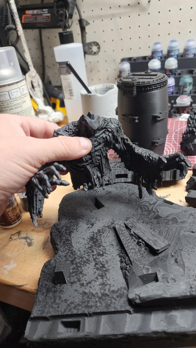 May include: A black 3D printed miniature figure of a monstrous creature with elongated arms and a textured, jagged body, positioned above a detailed, uneven terrain base featuring debris and rocky elements, all finished in a matte black primer.
