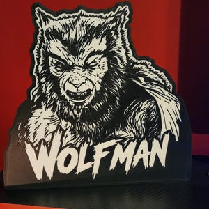 Peut inclure: Une plaque d&eacute;corative noire et blanche pr&eacute;sentant une illustration d&eacute;taill&eacute;e d'un loup-garou grognant avec une texture de fourrure et des dents pointues. Le mot WOLFMAN est imprim&eacute; en grandes lettres capitales blanches au bas de la pi&egrave;ce.