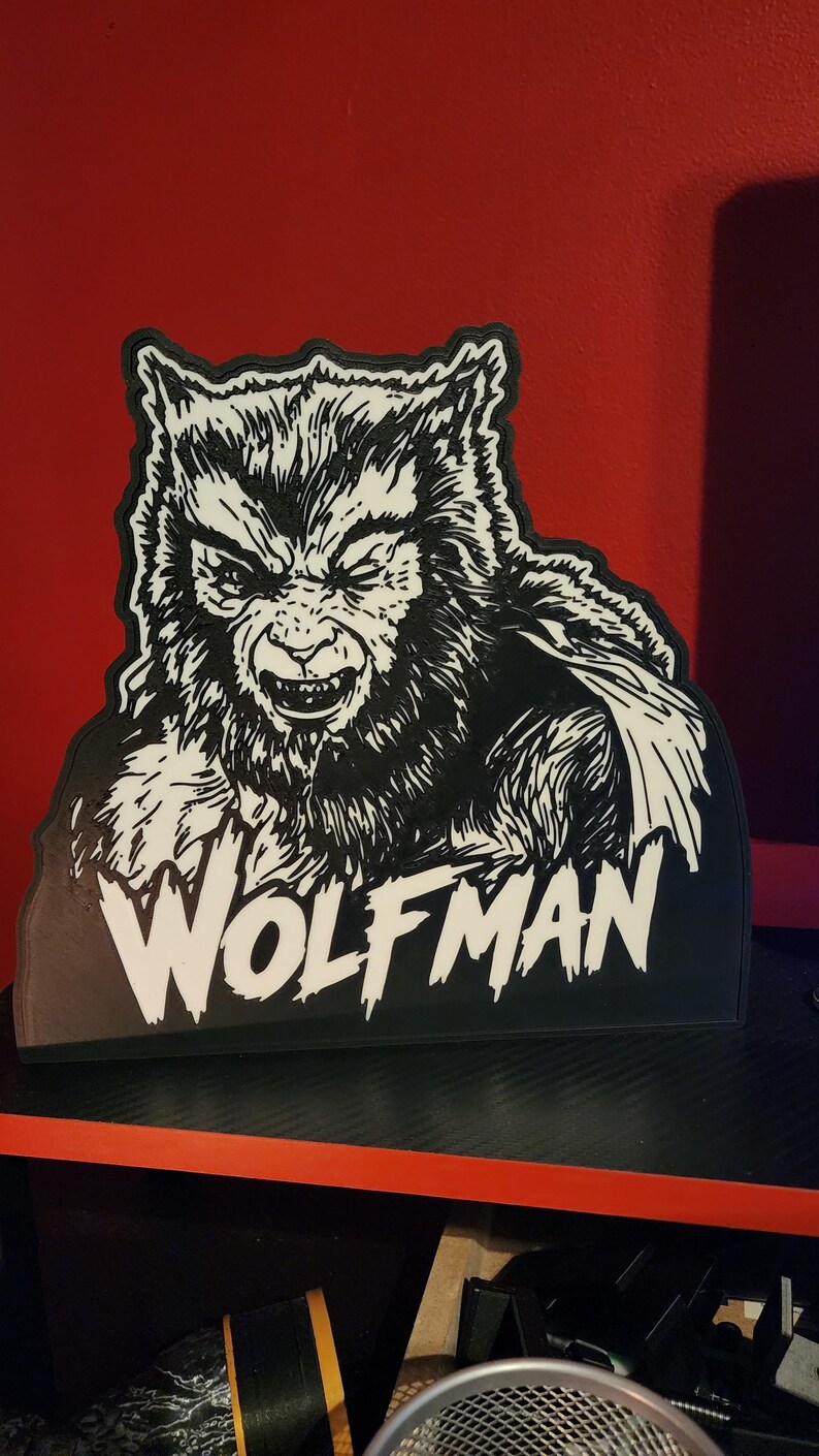 Peut inclure: Une plaque d&eacute;corative noire et blanche pr&eacute;sentant une illustration d&eacute;taill&eacute;e d'un loup-garou grognant avec une texture de fourrure et des dents pointues. Le mot WOLFMAN est imprim&eacute; en grandes lettres capitales blanches au bas de la pi&egrave;ce.