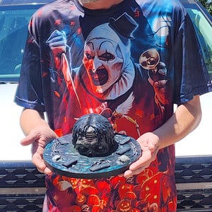 May include: A circular, dark-toned display base featuring a central sculpted head with dark hair, surrounded by small rectangular markers and chain links. The base has a textured, mottled blue and black surface finish, presenting a detailed horror-themed prop.