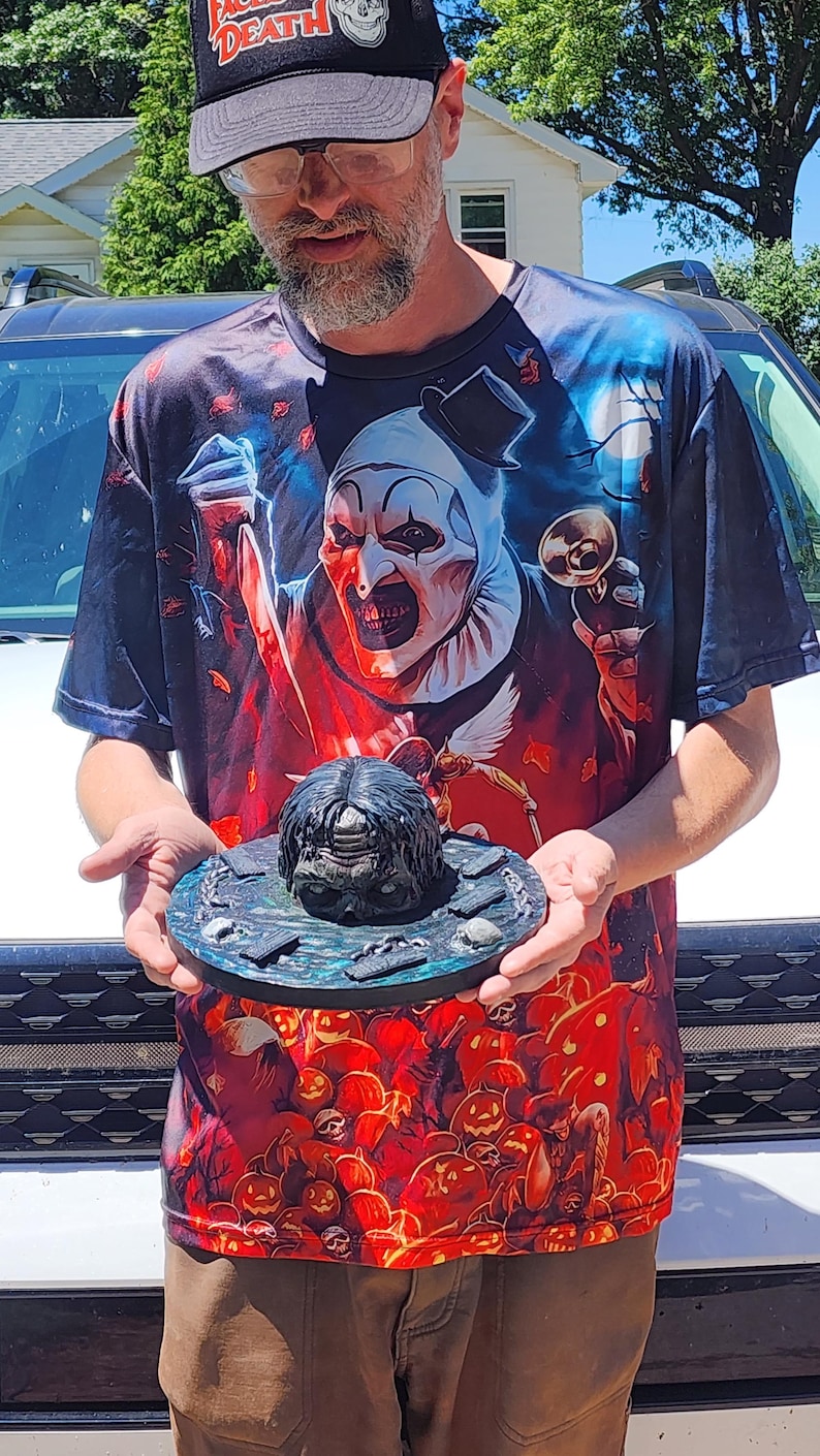 May include: A circular, dark-toned display base featuring a central sculpted head with dark hair, surrounded by small rectangular markers and chain links. The base has a textured, mottled blue and black surface finish, presenting a detailed horror-themed prop.