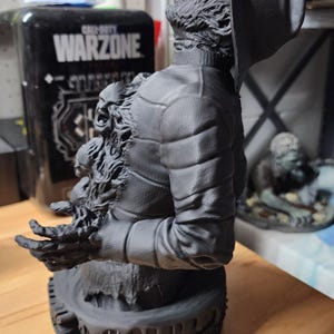 May include: A matte black 3D printed bust of a figure wearing a wide-brimmed hat and a quilted jacket. The figure features a textured, distressed appearance with a smaller, screaming face emerging from the chest area and outstretched fingers.