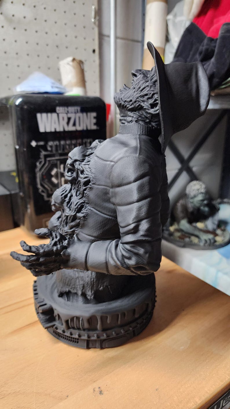 May include: A matte black 3D printed bust of a figure wearing a wide-brimmed hat and a quilted jacket. The figure features a textured, distressed appearance with a smaller, screaming face emerging from the chest area and outstretched fingers.
