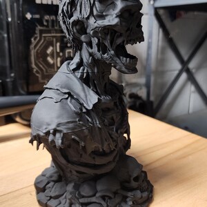 以下が含まれることがあります： 口を開け、歯が露出し、ぼろぼろの服を着た腐敗したゾンビの姿をした、マットブラックの3Dプリント製胸像。このフィギュアは、複数の人間の頭蓋骨と骨格の残骸で構成された台座の上に配置されており、複雑な質感が表現されています。