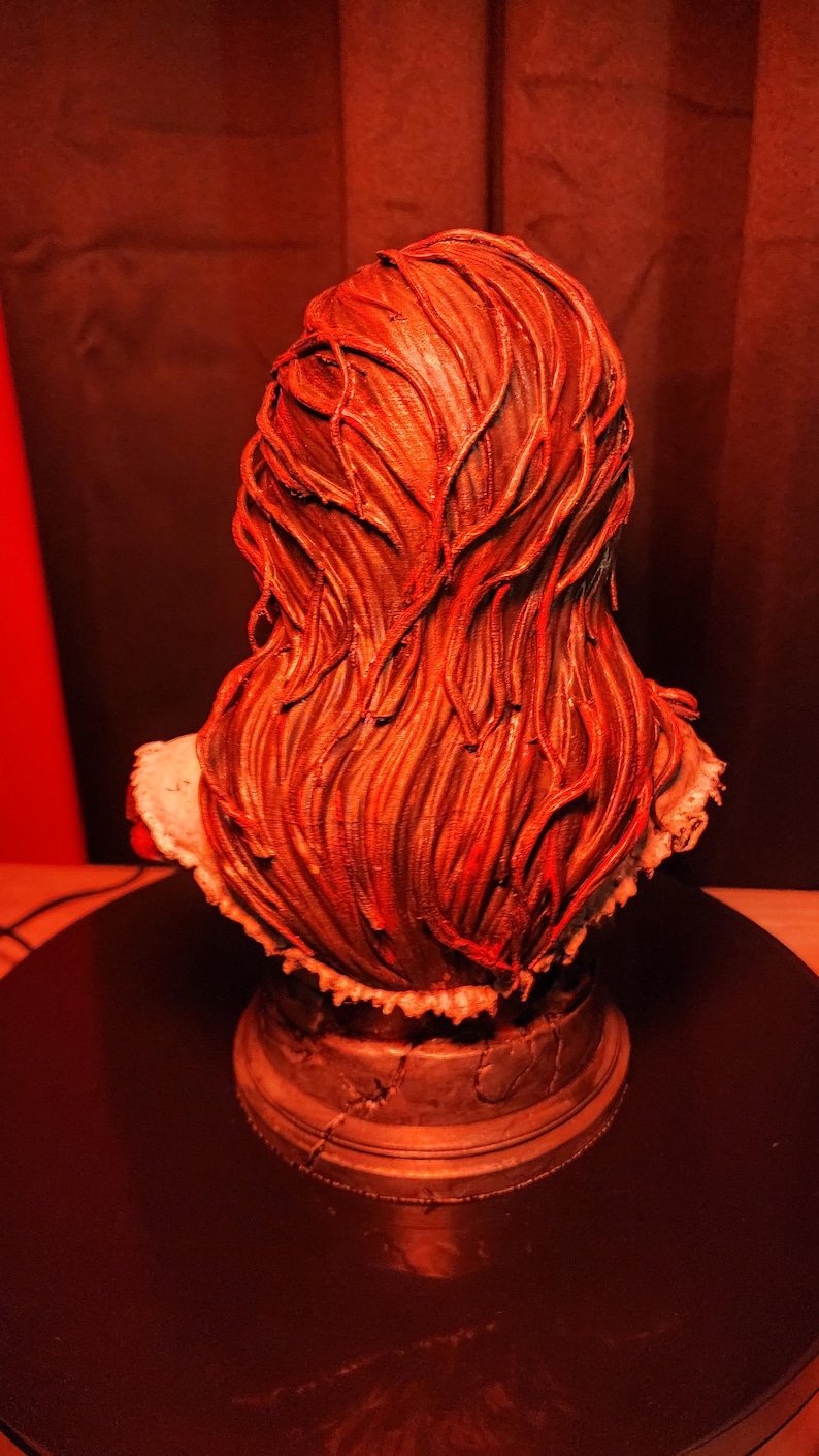 K&ouml;nnte beinhalten: Eine skulptierte B&uuml;ste einer Figur mit langem, flie&szlig;endem, strukturiertem Haar in tiefroten und braunen T&ouml;nen. Die Figur steht auf einem runden, verwitterten Sockel mit sichtbaren Rissen und wird von einem warmen roten Licht beleuchtet.