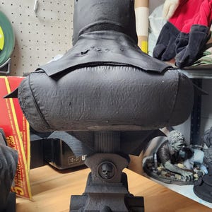May include: A matte black bust of a caped figure viewed from behind, featuring a textured surface. The figure rests on a pedestal base designed like a tombstone with a small skull emblem and a central pillar support, all in a matching black finish.