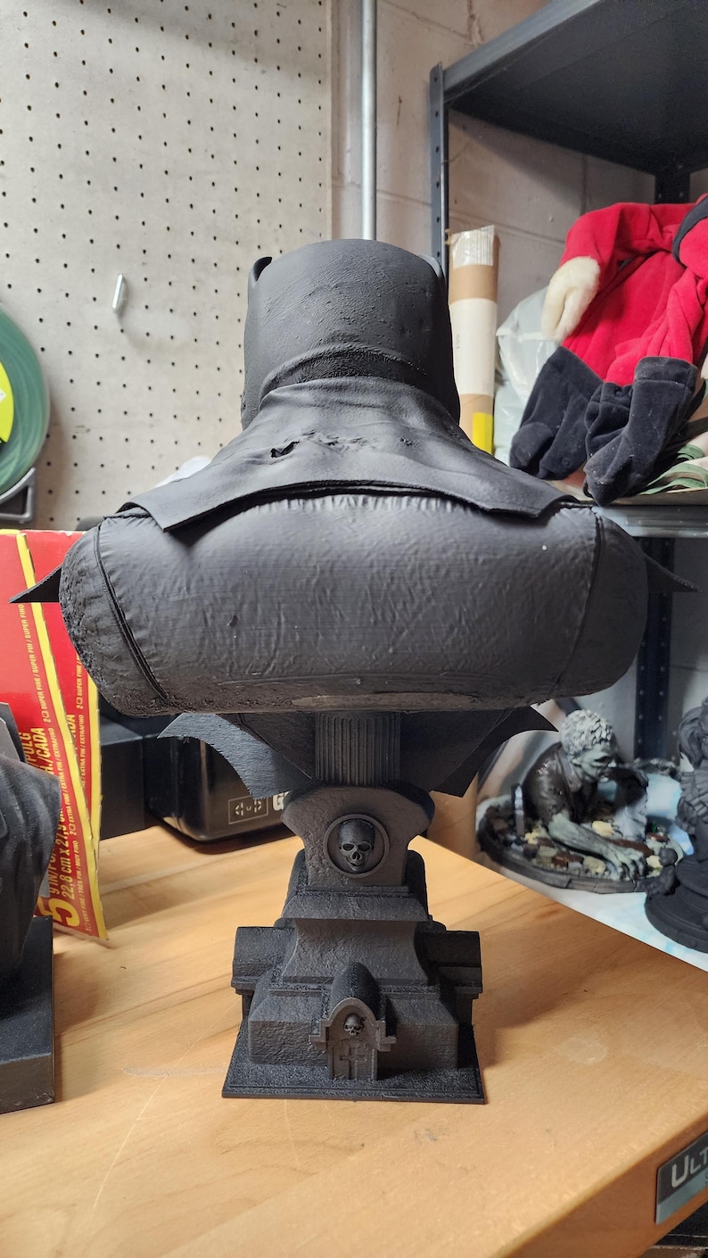 May include: A matte black bust of a caped figure viewed from behind, featuring a textured surface. The figure rests on a pedestal base designed like a tombstone with a small skull emblem and a central pillar support, all in a matching black finish.