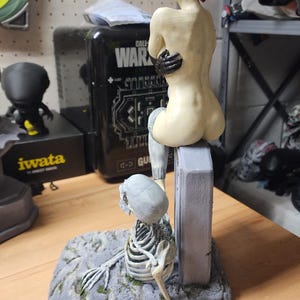 Puede incluir: Una figura pintada a mano que muestra a un personaje de piel p&aacute;lida con cabello corto rojo sentado sobre una l&aacute;pida gris. Una figura esquel&eacute;tica con cr&aacute;neo y caja tor&aacute;cica gris claro descansa en la base de la l&aacute;pida sobre un suelo rocoso gris.