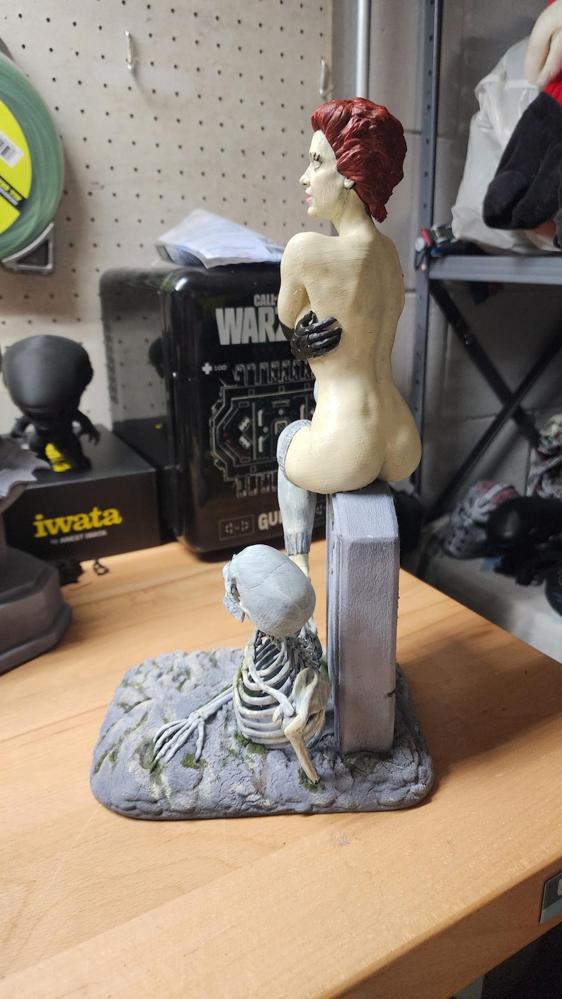 Puede incluir: Una figura pintada a mano que muestra a un personaje de piel p&aacute;lida con cabello corto rojo sentado sobre una l&aacute;pida gris. Una figura esquel&eacute;tica con cr&aacute;neo y caja tor&aacute;cica gris claro descansa en la base de la l&aacute;pida sobre un suelo rocoso gris.