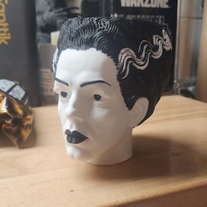 May include: A 3D printed planter shaped like the head of the Bride of Frankenstein, featuring a white face, black hair with a prominent white streak, black eyebrows, black eyelashes, and black lips, with a hollow top opening for plants.