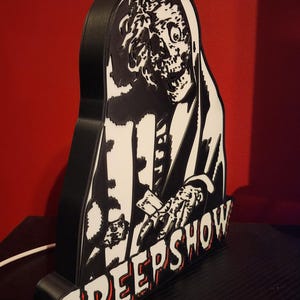May include: A black and white 3D printed sign featuring a skeletal figure with a hood and the text CREEPSHOW in bold, stylised lettering. The object has a thick black frame and a flat, graphic front panel depicting a detailed, high-contrast illustration.