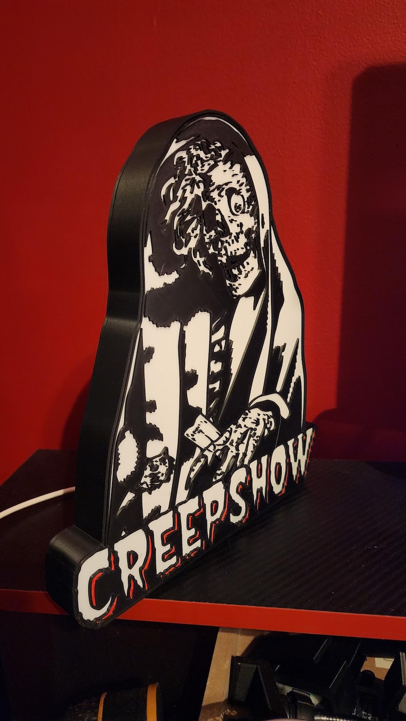 May include: A black and white 3D printed sign featuring a skeletal figure with a hood and the text CREEPSHOW in bold, stylised lettering. The object has a thick black frame and a flat, graphic front panel depicting a detailed, high-contrast illustration.