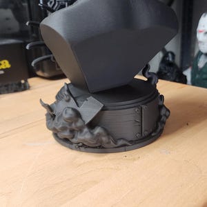 May include: A matte black bust of a figure wearing a wide-brimmed hat, positioned on a circular base decorated with sculpted smoke and a small axe. The sculpture features a textured collar and a smooth, solid black finish throughout the piece.