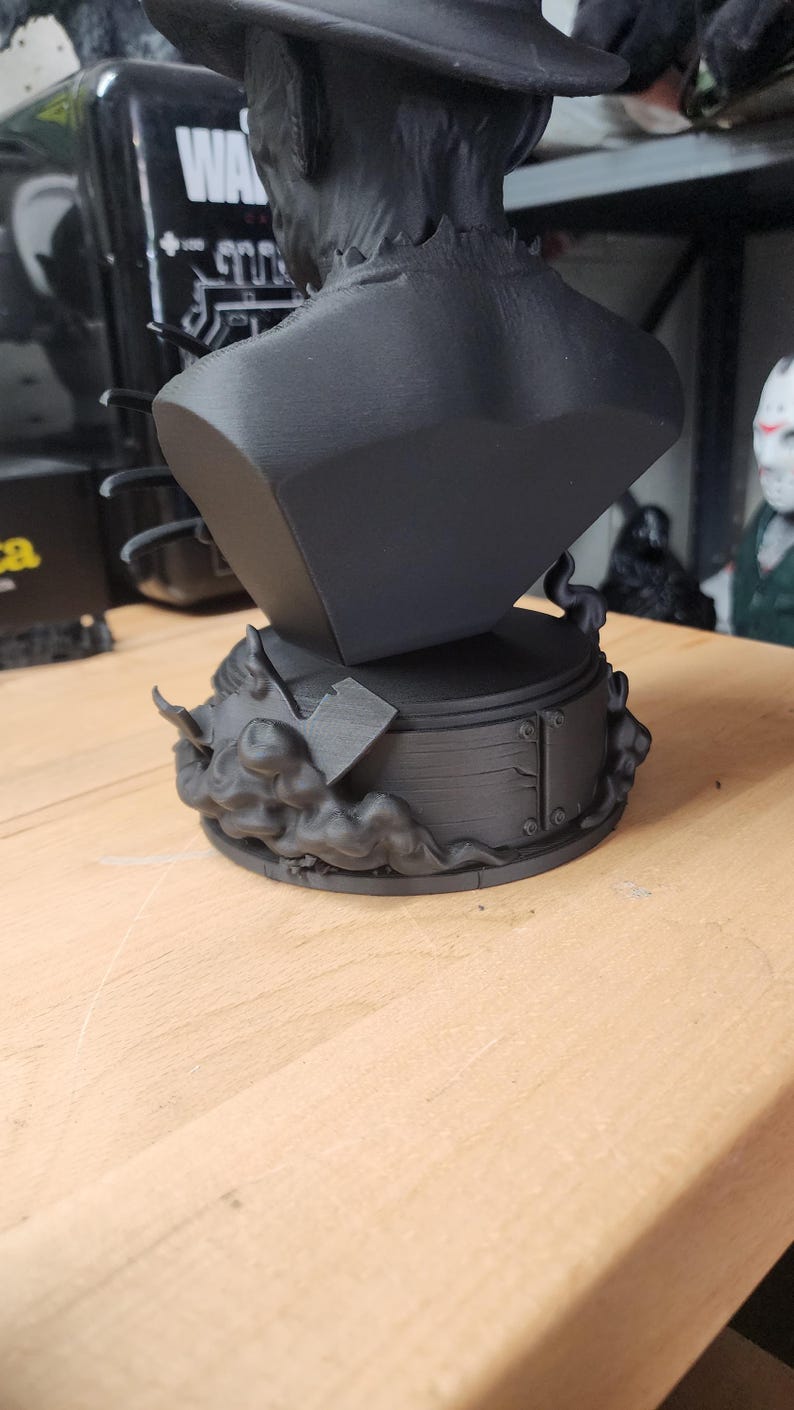 May include: A matte black bust of a figure wearing a wide-brimmed hat, positioned on a circular base decorated with sculpted smoke and a small axe. The sculpture features a textured collar and a smooth, solid black finish throughout the piece.